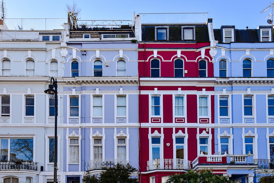 A row of three-story Victorian-style terraced houses painted in pastel shades, with the middle building highlighted in a vibrant red and the adjacent homes in light blue and white. The facades feature ornate decorative molding and window frames, with rectangular sash windows on each floor. The rooftops are sloped with dark tiles, and dormer windows are visible on the top levels. A black vintage-style lamppost stands in the foreground on a paved sidewalk, partially obscuring the lower part of the red building. The lower windows have curtains drawn, and small balconies with white wrought-iron railings are attached to some of the upper-facing windows. The scene is lit with natural daylight, and the background shows a clear blue sky, with no visible rubbish or waste in the environment, suggesting a clean, residential area connected to private property management or alternative waste collection methods. The image is clear and sharply focused, emphasizing the architectural details and color contrast of the row houses, relevant for content about property appearance and external features of residential rubbish removal services in urban settings.