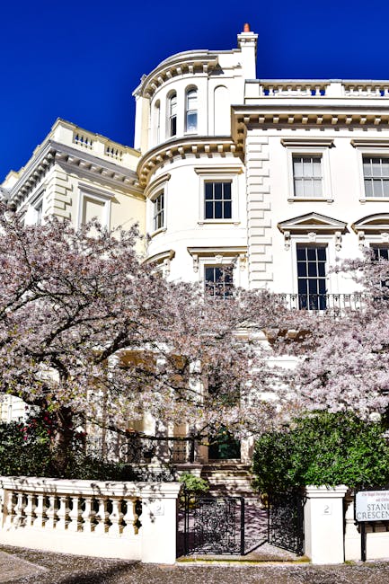 The image depicts a large, elegant white Victorian-style building with ornate architectural details, including rounded bay windows and decorative cornices, set against a vivid blue sky. In front of the building, there are blooming pink and white cherry blossom trees with dense clusters of flowers, partially obscuring the lower part of the structure. The property features a small front garden area with green shrubbery and a wrought iron gate situated between stone pillars. A low cream-colored balustrade runs along the edge of the garden, and a pathway leading to the entrance is partially visible through the trees. The scene is well-lit with natural daylight, highlighting the contrast between the white facade, the pink blossoms, and the deep blue sky, illustrating a typical residential setting that may require occasional private rubbish clearance services from companies like rubbishclearancenottinghill.com for maintaining cleanliness around such high-value properties.