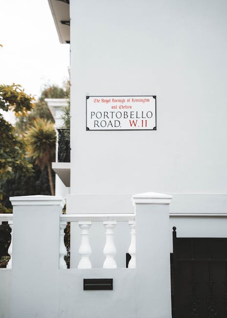 A close-up view of a white residential building featuring a small front garden enclosed by a low wall with decorative balustrades. On the wall, there is a rectangular street name sign reading 'Portobello Road W.11' with a statement indicating it is part of the Royal Borough of Kensington and Chelsea. The building's exterior wall is smooth and painted white, with minimal architectural detail visible. To the left, a small section of an upper balcony with a white railing can be seen, alongside mature trees and shrubs with green and yellow foliage in the background, indicating a suburban or urban residential setting. The lighting suggests natural daylight with a soft, even tone, and the scene appears clean and well-maintained. This image subtly relates to private property and independent disposal options for rubbish removal, as indicated by the context of alternative waste handling in local residential areas, supported by the clean and organized appearance of the site that is consistent with professional rubbish clearance services.