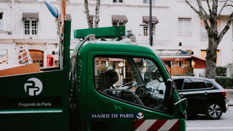 A close-up view of a green waste collection truck operated by the Mairie de Paris, parked on a residential street with multi-storey buildings and leafless trees in the background. The truck's cab features a transparent side window revealing the interior with a visible steering wheel and dashboard. Mounted on the top of the truck is a hydraulic lift mechanism, likely used for lifting and emptying waste containers. The side panel displays a white logo and the words 'Propreté de Paris' in white text, indicating municipal waste services. A black car is parked behind the truck, and a building with large windows, an awning, and outdoor furniture can be seen further in the background. The scene is lit by natural daylight, with shadows cast on the pavement, illustrating a typical urban environment where private waste collection and rubbish removal services may complement or provide alternatives to local authority refuse collection methods.