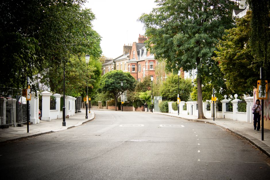 A close-up view of a white residential building featuring a small front garden enclosed by a low wall with decorative balustrades. On the wall, there is a rectangular street name sign reading 'Portobello Road W.11' with a statement indicating it is part of the Royal Borough of Kensington and Chelsea. The building's exterior wall is smooth and painted white, with minimal architectural detail visible. To the left, a small section of an upper balcony with a white railing can be seen, alongside mature trees and shrubs with green and yellow foliage in the background, indicating a suburban or urban residential setting. The lighting suggests natural daylight with a soft, even tone, and the scene appears clean and well-maintained. This image subtly relates to private property and independent disposal options for rubbish removal, as indicated by the context of alternative waste handling in local residential areas, supported by the clean and organized appearance of the site that is consistent with professional rubbish clearance services.