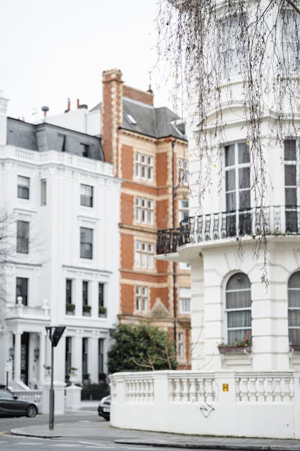 The image depicts a street scene in a residential area, featuring a white, multi-story building with classic architectural details, including a rounded corner tower with decorative railings on a small balcony. The building's facade has arched windows with window boxes containing small plants or flowers. To the left, partially visible, is another white building with a modern design, and further back, a taller red-brick building with contrasting lighter accents and traditional detailing. A tree with bare branches overhangs the scene, indicating a season such as late autumn or winter. The street appears clean, with a few parked cars visible along the curb, and a streetlamp standing in front of the buildings. The overall setting suggests an urban residential neighborhood, common in areas where private rubbish collection or on-site clearance services like those provided by rubbishclearancenottinghill.com might be utilized for managing waste and rubbish removal from properties with period architecture and limited street-level storage space.