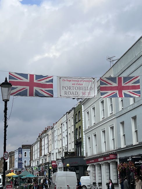 A street scene on Portobello Road W11 featuring a row of terraced buildings painted in white, light green, and dark gray in a typical urban residential style. The buildings have multiple floors with uniformly arranged windows, some with black wrought-iron balconies. In the foreground, parked white vans are visible along the street lined with various small shops and vendors; some with awnings and outdoor displays. Overhead, a large banner displaying the Union Jack flag and reading 'The Royal Borough of Kensington and Chelsea - Portobello Road W11' spans across the street, supported by cables attached to lampposts. The lamppost on the left side has a traditional design with ornate black metalwork, and the sky above is overcast with thick gray clouds, creating diffuse lighting that highlights the textures of the building façades and street scene. The overall scene captures an active, typical day on this well-known street, which often involves private rubbish collection and local waste management services, consistent with alternative disposal options outside municipal collection.