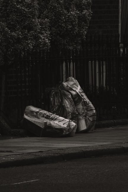 Two large black rubbish bags made of durable plastic, tightly knotted at the top, are placed on the edge of a paved sidewalk near a black metal fence with vertical bars and a trellis panel behind. The bags appear filled with household waste and are positioned side by side, leaning slightly against each other. In the background, dense foliage from a tree or shrub extends upward, partially obscuring some of the fence. The scene is outdoors in a residential area, with the street surface visible in the foreground. The lighting suggests overcast conditions, casting minimal shadows. The presence of the bags and the surrounding environment indicate an informal waste disposal area, reflecting scenarios where private rubbish collection services or on-site clearance might be employed, in contrast with municipal waste collection policies.