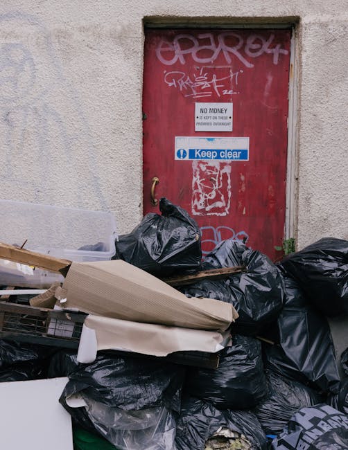 A collection of black rubbish bags, some torn open, is piled in front of a red metal door with multiple layers of graffiti and stickers, including a white sticker with the text 'NO MONEY' and a blue 'Keep clear' sign. The rubbish bags are spilling over a makeshift arrangement of cardboard and wooden debris, suggesting an on-site or private collection of waste outside a building with rough, beige exterior walls. The scene depicts an informal disposal area, likely part of a non-recycling waste clearance, with waste materials positioned on a pavement or outside a rear alleyway, indicating a situation where external waste removal services or private disposal methods are in use, consistent with policy considerations around household waste management and alternative rubbish handling.
