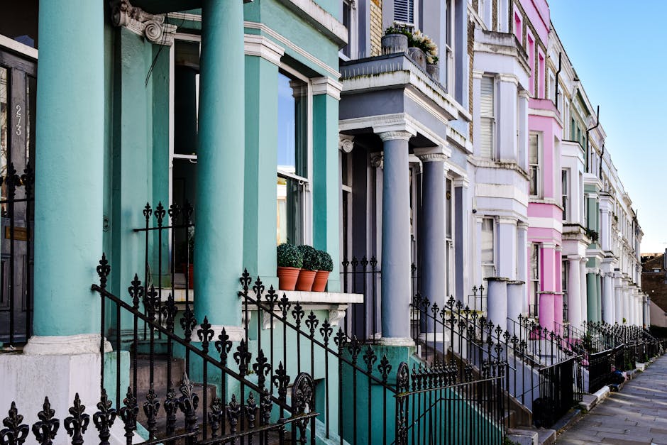 The image depicts a row of Victorian-style terraced houses on a city street, each featuring front steps leading up to small porches with ornate black wrought iron railings. The facades are painted in pastel shades including teal, lavender, pink, and mint green, with the colors contrasting against white window frames and decorative mouldings. Large, rounded columns support porches on the teal and lavender houses, while potted plants with green, rounded foliage sit on some of the window sills. The street is quiet, with the pavement visible in the foreground and a clear blue sky in the background, creating a neat, orderly urban scene. The setting suggests a typical residential area where private waste collection or on-site clearance services, like those provided by rubbish clearance specialists such as rubbishclearancenottinghill.com, may be used to manage waste left outside the properties, especially in contexts where avoiding local authority fly-tipping is a concern.