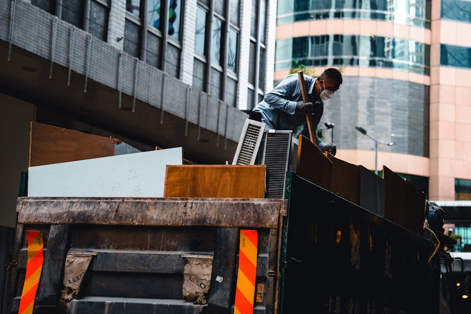 A man wearing a white face mask, a light blue shirt, and dark trousers is standing on the edge of a large, open-top waste truck, leaning forward as he loads various types of discarded furniture and wooden panels into the truck. The furniture includes a brown wooden cabinet, a white panel, and a dark-colored vent or air conditioning unit, all with visible textures and finishes such as smooth surfaces and grain patterns. The background features a modern urban environment with high-rise glass and concrete buildings, some with reflective windows, and a concrete staircase or bridge overhead. The truck's bed appears weathered, with rust and dirt visible on its sides, suggesting an active rubbish removal service. The scene is illuminated naturally, with soft lighting-enhanced by overcast sky conditions, conveying a professional and neutral tone relevant to private waste handling or alternative rubbish collection services in a city setting. The overall composition highlights on-site clearance activities focused on bulky waste disposal, aligning with the context of independent rubbish removal solutions offered by companies like rubbishclearancenottinghill.com.