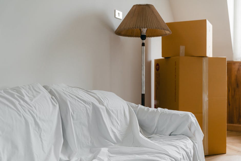 In a room with neutral-colored walls and wooden flooring, three cardboard boxes of varying sizes are stacked against the wall, positioned near a wooden cabinet. The boxes are sealed with packing tape and appear to be used for moving or storage. To the left, a white fabric sofa with slightly rumpled upholstery occupies part of the space, with no visible decorative cushions. Adjacent to the boxes, a standing floor lamp with a metallic pole and a large, pleated beige lampshade illuminates the area, casting soft light. The environment looks tidy and organized, suggesting a typical interior setting where furniture and packing materials are temporarily stored or prepared for disposal. The scene subtly reflects aspects of private waste handling, characteristic of alternative rubbish removal services by [COMPANY_NAME], as part of an indoor clearance or pre-move rubbish collection process.