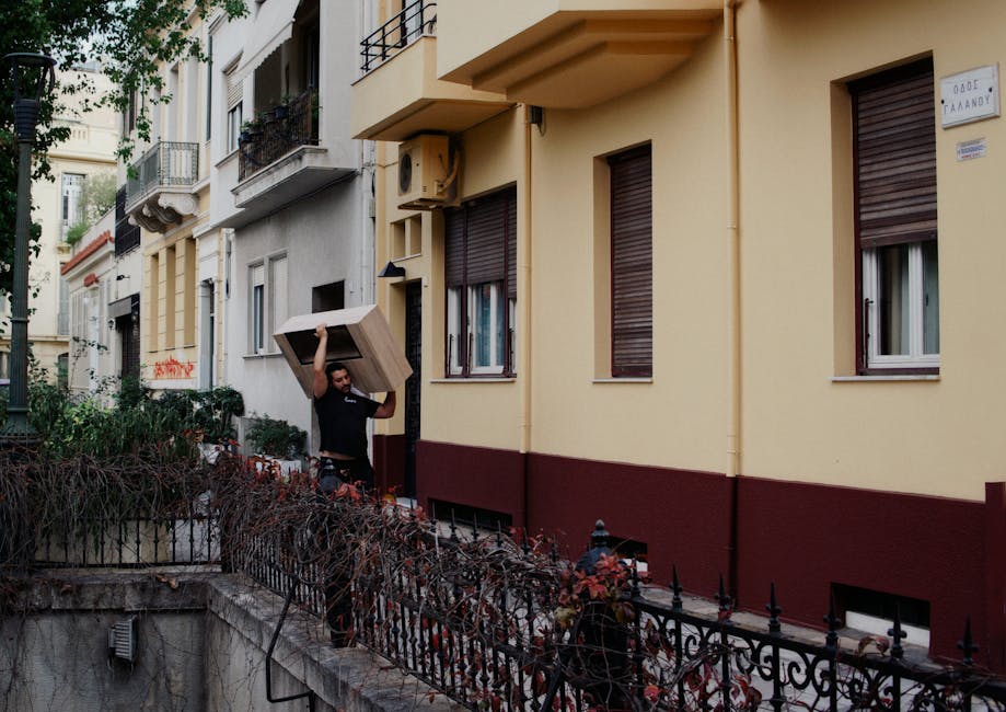 A man carrying a medium-sized wooden cabinet, which appears to be a piece of furniture with a smooth, polished light brown finish, is walking along a narrow urban sidewalk adjacent to a residential building. The cabinet is being held upright with one hand on its top edge, and the man is dressed casually in a dark t-shirt and patterned shorts. The building behind him features a yellow façade with three closed brown window shutters, one of which has a small curtain visible behind it. To the right, an external air conditioning unit is mounted on the wall, alongside a small wall-mounted sign. The sidewalk is bordered by a low, black metal fence with decorative finials, which surrounds a small front garden filled with dried and dormant plants. The scene is lit with soft natural daylight, indicating daytime, and the environment suggests a typical residential street in Notting Hill with multi-storey apartments. This image relates to private rubbish or furniture removal, often handled by independent waste collection services such as rubbishclearancenottinghill.com, demonstrating the process of on-site clearance of bulky items.