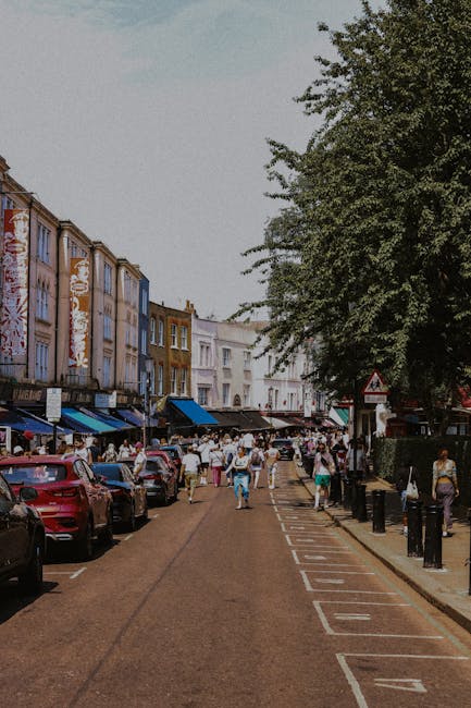 A busy urban street scene in Notting Hill with a row of multi-story buildings featuring colorful facades, some with visible brickwork and large windows. The street is lined with parked cars on both sides, including a red vehicle in the foreground, and has designated parking bays marked along the edge. Pedestrians populate the pavement, some walking and others browsing outdoor market stalls with blue awnings, suggesting a marketplace or shopping area. To the right, there is a large leafy tree providing shade, with black bollards along the sidewalk to manage foot traffic. The atmosphere appears lively and well-lit under natural daylight, highlighting the typical aesthetic of a vibrant neighborhood. Although not directly related to rubbish removal, the image may evoke the importance of efficient waste disposal for maintaining cleanliness in busy commercial and residential zones like Notting Hill, where private rubbish collection and on-site clearance services by companies such as rubbishclearancenottinghill.com are often used to complement public waste management efforts.