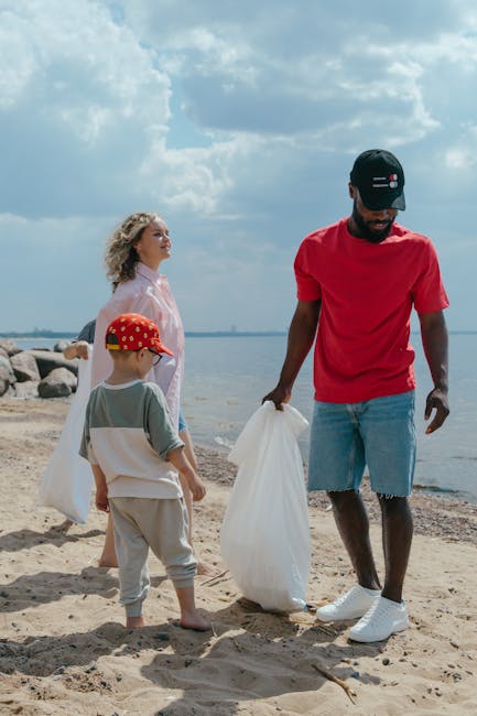 A man wearing a red T-shirt, black cap, and light blue denim shorts stands on a sandy beach, holding a white plastic trash bag in one hand and inspecting a second bag. To his left, a young boy in a light gray T-shirt, beige pants, and a red cap with yellow dots also holds a clear plastic bag, facing the water. Behind them, a woman with curly blonde hair, dressed in a light pink or white long-sleeve top, looks out over the shoreline. The scene features a calm body of water with a rocky jetty extending into it, and a partly cloudy sky overhead, suggesting a clear, bright day. The setting implies a shoreline cleanup effort, with the individuals collecting rubbish to support environmental preservation, aligning with the context of independent waste collection or on-site clearance, natural to private rubbish removal services such as those offered by companies like rubbishclearancenottinghill.com.