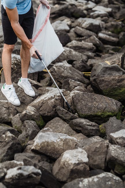 A person wearing a light blue t-shirt, black shorts, and white sneakers is using a litter picker or grabber tool to collect scattered debris from a rocky shoreline or riverbank. The individual is holding a large white trash bag to contain the collected rubbish, focused on clearing the uneven area covered with various-sized stones and pebbles that have a mix of dark grey, brown, and mossy tones. The environment appears outdoors with natural lighting, highlighting the textured surfaces of the rocks and the informal setting. This scene showcases a commitment to environmental cleanliness and might relate to private waste collection or disposal efforts by services such as rubbishclearancenottinghill.com, emphasizing independent or on-site rubbish removal practices rather than municipal collection, aligned with the context of alternative waste handling methods.