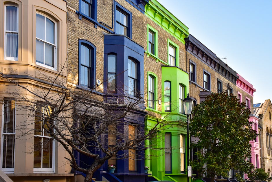 The image features a row of multi-story Victorian-style terraced buildings with colorful facades, each exhibiting distinct painted finishes. From the left, one building has a beige exterior with tall, white-framed sash windows and ornate window surrounds. Adjacent to it is a structure painted in deep navy blue, with similarly styled windows and decorative molding. Next, a building is painted bright green, with matching trim highlighting its bay window and cornice. To the right, another building displays a muted yellow brick facade with darker window frames, followed by a pink-painted building with similar architectural details. In the foreground, a leafless tree with thin, winding branches partially obstructs the view, and a traditional black streetlamp stands near the green building. The scene is illuminated by daylight against a clear, blue sky, emphasizing the vibrancy of the painted exteriors while the environment suggests an urban setting with well-maintained residential buildings. This scene subtly aligns with the context of independent waste and rubbish handling suitable for private property or on-site clearance, as often handled by local rubbish removal services like rubbishclearancenottinghill.com.