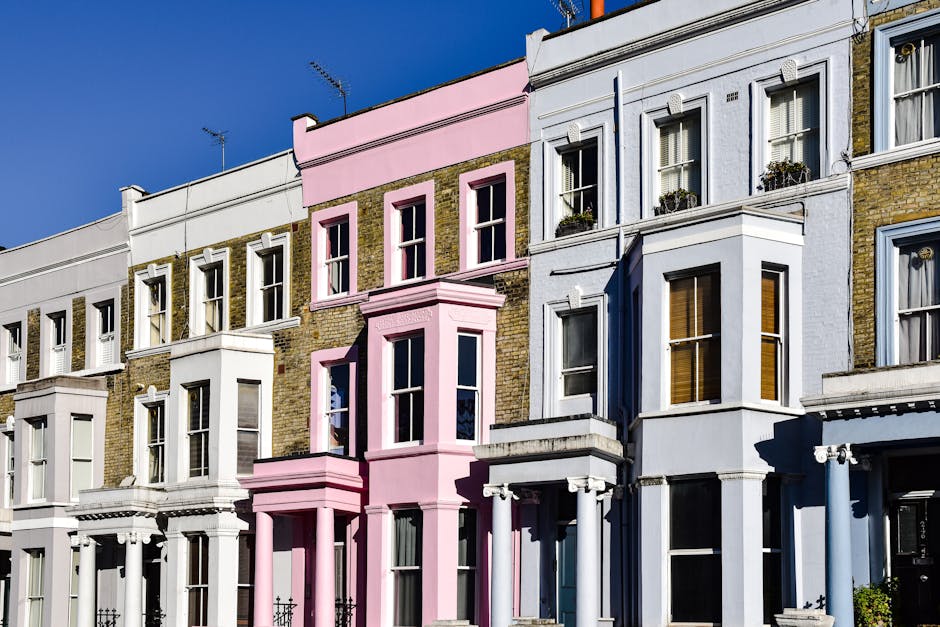 Three adjacent Victorian-style residential buildings with distinct architectural features are visible in this image. The building on the left features a white façade with decorative moldings, large sash windows, and small balconies with railing details. The central building has a striking pink exterior with a cornice at the roofline, tall narrow windows framed in white, and a protruding bay window on the second floor supported by decorative brackets, all set against exposed brickwork that adds texture. The rightmost building is painted in light blue with similar sash windows, some of which have window boxes filled with plants. The façades are finished with smooth stucco and painted woodwork, creating a contrast with the brickwork on the pink building. The scene is illuminated by bright, natural daylight, with shadows cast by the architectural elements, set against a clear blue sky. The image showcases typical urban residential architecture, with no visible rubbish or outdoor waste, fitting into a context where independent waste handling or private rubbish removal services could be relevant.