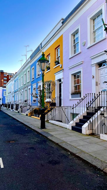 The image features a row of multi-story Victorian-style terraced buildings with colorful facades, each exhibiting distinct painted finishes. From the left, one building has a beige exterior with tall, white-framed sash windows and ornate window surrounds. Adjacent to it is a structure painted in deep navy blue, with similarly styled windows and decorative molding. Next, a building is painted bright green, with matching trim highlighting its bay window and cornice. To the right, another building displays a muted yellow brick facade with darker window frames, followed by a pink-painted building with similar architectural details. In the foreground, a leafless tree with thin, winding branches partially obstructs the view, and a traditional black streetlamp stands near the green building. The scene is illuminated by daylight against a clear, blue sky, emphasizing the vibrancy of the painted exteriors while the environment suggests an urban setting with well-maintained residential buildings. This scene subtly aligns with the context of independent waste and rubbish handling suitable for private property or on-site clearance, as often handled by local rubbish removal services like rubbishclearancenottinghill.com.
