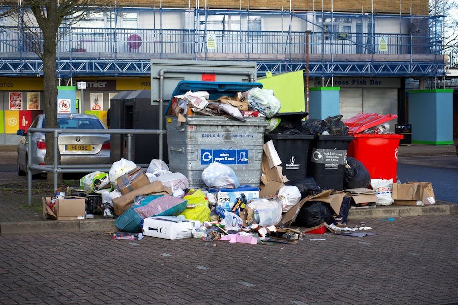 In the foreground of the image, a large pile of mixed household waste and recyclables is carelessly accumulated on a paved sidewalk area. The rubbish includes cardboard boxes, plastic bags, sealed black garbage bags, and various paper and packaging materials, some spilling onto the surrounding brick pavement. Prominently at the center, there is a large grey mixed waste bin overflowing with loose paper and cardboard, with its lid partially open revealing the contents. To the right of this bin, black and red recycling bins are visible, also filled with waste, with plastic bags and other refuse stacked nearby. On the left, a parked silver car is situated behind a low metal barrier, with a tree positioned behind the vehicle, its trunk partially visible. The background features a commercial building with a yellow storefront and a blue metal-framed balcony or scaffold, with signs indicating different businesses and safety notices. The overall scene suggests an area receiving a private or alternative rubbish clearance, possibly in preparation for scheduled collection or removal by a waste management service such as rubbishclearancenottinghill.com, with an urban environment typical of a residential or retail frontage affected by improper disposal or delayed refuse collection.