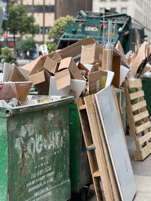A large green wheeled waste bin is filled with a variety of discarded cardboard boxes, some flattened and others still in their original shape, along with white packaging inserts and smaller packaging materials. The cardboard boxes are primarily brown, with some having printed labels or markings visible on the sides. The bin is situated on an urban street, with a backdrop of trees and multi-story buildings, and is positioned near a green waste collection vehicle and wooden pallets that lean against the bin's side. The scene suggests an on-site clearance of rubbish, typical of independent waste disposal processes that may be arranged through private waste removal services, such as those provided by Rubbish Clearance Notting Hill. The environment appears well-lit, with natural daylight illuminating the scene, highlighting the textures of the cardboard and the weathered surface of the bin, which shows some signs of dirt and usage typical of waste collection points in busy city streets. The overall composition emphasizes the practical aspects of rubbish removal and the volume of waste generated in such waste management activities.