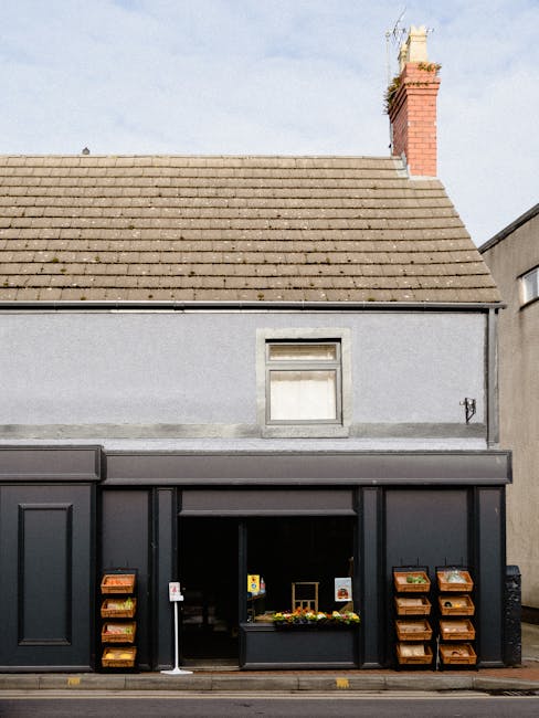 The image depicts a two-story building with a sloped tiled roof and a prominent red brick chimney stack extending above the roofline. The upper floor features a small, rectangular window with a white frame and a partially closed blind, set into a smooth, light grey exterior wall. The ground floor comprises a black-painted shopfront or commercial space with large glass windows and a centrally positioned entrance. To the left and right of the entrance are angled wooden display racks affixed to the black façade, each containing multiple compartments filled with various small items, possibly plants or packaging materials. The building is situated on a city street with minimal surrounding context visible. The natural daylight illuminates the scene, revealing the textures of the tiled roofing, smooth painted walls, and the wooden racks. Overall, this setup suggests a retail or small business space that might involve occasional private waste collection or on-site clearance, consistent with the context of independent rubbish removal services by [COMPANY_NAME], as observed in typical commercial premises. This scene emphasizes the practical arrangement of a commercial property, positioned between blank wall sections and accessed directly from the pavement.